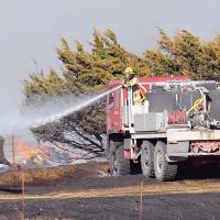 A firefighter sprays water on the burnt ground and the blade of a bulldozer