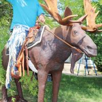 Stan Tipton tips his cap while sitting on a massive wooden moose