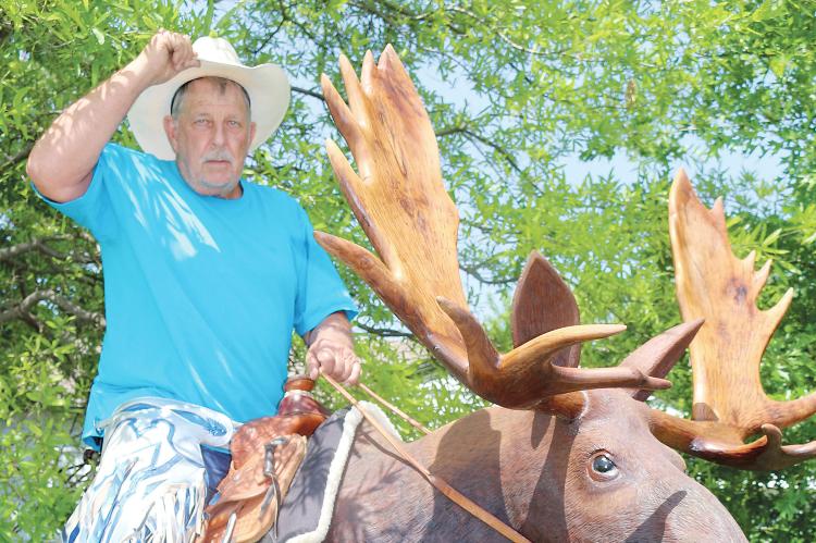 Stan Tipton tips his cap while sitting on a massive wooden moose